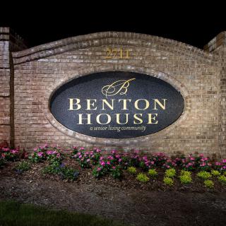A well-lit Benton House sign is displayed on a brick wall surrounded by flowers at night.