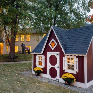 A charming red and white playhouse sits in the yard, surrounded by trees and flowers, with a larger home visible in the background during the evening.