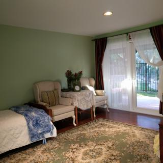 A cozy bedroom with a bed, two armchairs, and a rug, illuminated by natural light through sheer curtains.