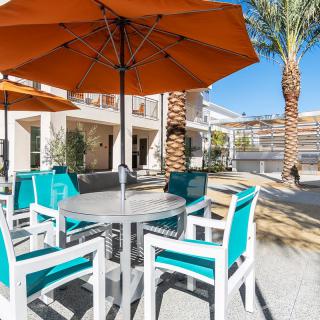 A sunny outdoor patio area with tables, chairs, and large umbrellas surrounded by palm trees.