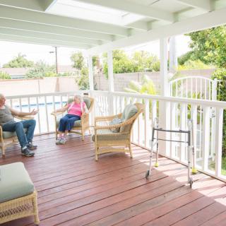 Elderly couple sitting on a porch with wicker furniture and a walker nearby.