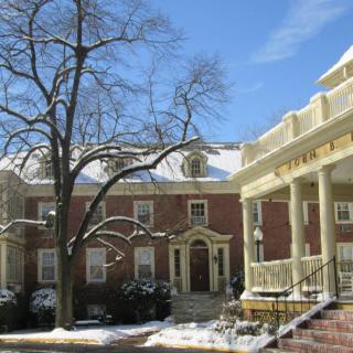 A brick building with columns, surrounded by snow and a large tree in winter sunlight.