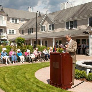 A speaker addresses an audience seated outdoors in front of a building.