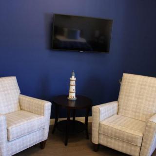 Two patterned chairs beside a small side table with a lighthouse ornament, situated under a wall-mounted television against a blue wall.