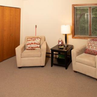 A cozy living room with two beige chairs, a small side table with a lamp, and a window.