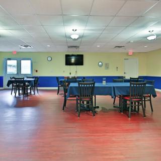 An empty dining room with several tables and chairs arranged, featuring a wooden floor and a wall-mounted television.