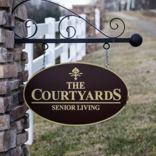 A metal sign for 'The Courtyards Senior Living' displayed outdoors next to a brick pillar and wooden fence.