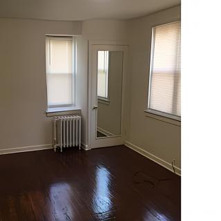 An empty room with hardwood flooring, featuring two windows, a radiator, and a door with a mirror.
