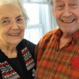 An elderly couple smiling indoors, standing near a window.