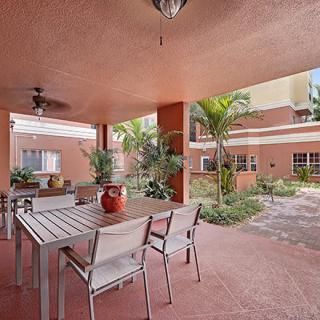A covered outdoor patio with a table and chairs, surrounded by plants and part of a building's exterior.