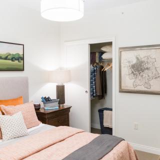 A neatly organized bedroom featuring a bed with orange and gray bedding, an open closet, and framed artwork on the walls.