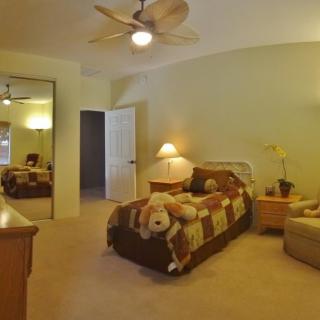 A cozy bedroom with a neatly made bed adorned with a teddy bear, a ceiling fan overhead, and a mirrored closet door reflecting the room.