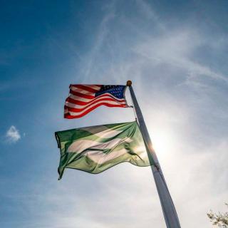 American and Nigerian flags flying on a pole against a sunny sky.