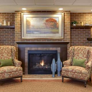 A cozy living room with a fireplace flanked by two patterned armchairs, a brick wall, decorative vases, and a painting above the mantel.