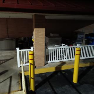 A dimly lit parking lot featuring a white fence and yellow safety poles near a building at night.