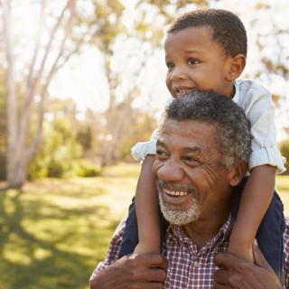 A grandfather carries his smiling grandson on his shoulders in a sunny park.
