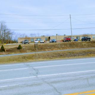 A view of a road lined with vehicles, leading to a building in the background under an overcast sky.