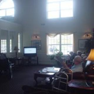A cozy living room with people sitting in chairs, a television, and large windows.