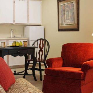 A cozy living space with a red armchair, a small table and chair, and a kitchenette in the background.