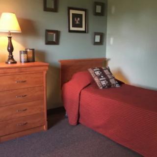 A cozy bedroom with a single bed and wooden dresser, warmly lit by a lamp.