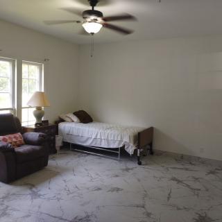 A minimalist bedroom with a single bed, armchair, ceiling fan, and natural light coming through the windows.