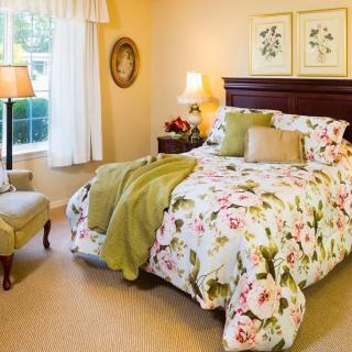 A cozy bedroom featuring a bed with floral-patterned bedding and decorative pillows, complemented by a warm lamp light and cushioned chair by the window.