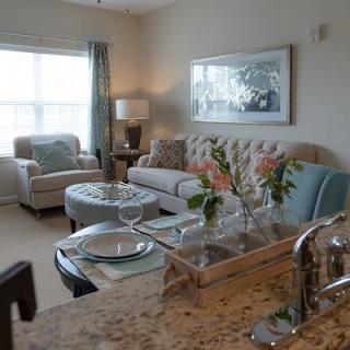 A cozy and elegantly decorated living room with a sofa, armchair, and coffee table adorned with flowers. Natural light streams in through a large window.