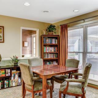 A cozy reading room with a wooden table, chairs, and a well-stocked bookshelf near large windows.