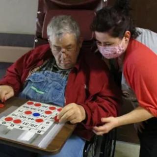 An elderly person is playing bingo with assistance from a caregiver wearing a mask.