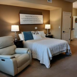 A cozy bedroom featuring a neatly made bed with decorative pillows, flanked by nightstands and lamps, alongside a comfortable recliner chair.