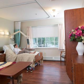 A hospital room with a patient in bed, featuring a large window and a vase of flowers.