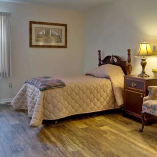 A cozy bedroom featuring a single bed with a wooden nightstand and lamp, an armchair, and a framed picture on the wall.