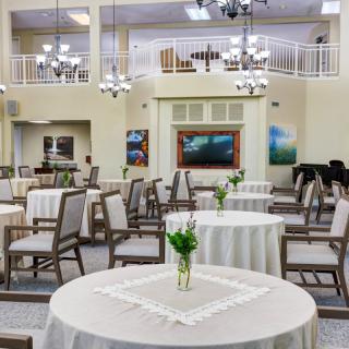 A spacious and elegantly decorated dining area featuring round tables set with white cloths and simple centerpieces, under subtle chandelier lighting.