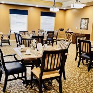 A well-lit dining room with neatly arranged tables and chairs, featuring warm decor and carpeted flooring.