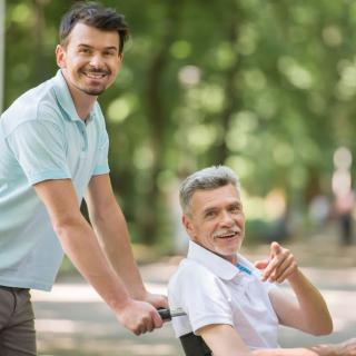 A caregiver pushing an elderly man in a wheelchair through a park.