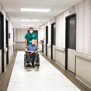 A healthcare worker assists a patient in a wheelchair down a hospital hallway.