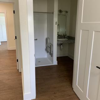 A view of a bathroom with a shower and sink, featuring white walls and wood flooring.