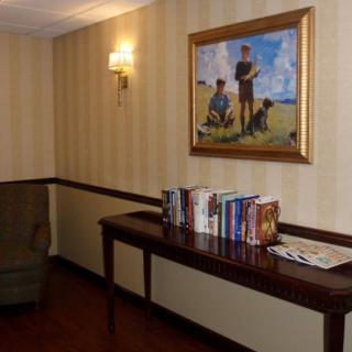 A cozy corner with a wooden table displaying books beneath a framed painting, illuminated by a wall sconce against striped wallpaper.