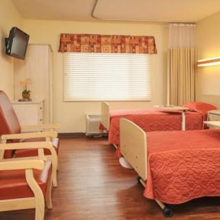 A clean and well-organized hospital room featuring two patient beds, chairs, a television, and a window with curtains.