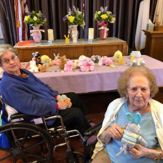 Two elderly women sitting indoors with stuffed animals and flowers on display.