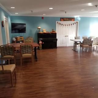 A spacious room with wooden flooring, featuring multiple tables and chairs, a piano, and a wall-mounted television.