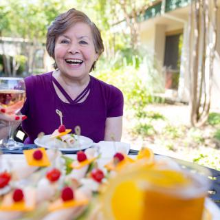 A woman smiling with a glass of wine, enjoying lunch outdoors in a garden setting.
