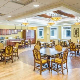A well-lit dining area with wooden tables and chairs, featuring chandeliers and an open kitchen space.