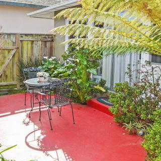 A cozy patio with a red floor, surrounded by lush plants and featuring a small outdoor table and chairs.