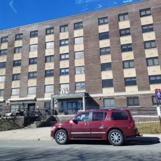 A red car parked in front of a multistory brick apartment building.