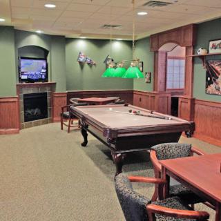 A cozy billiard room with a pool table, green walls, and wood paneling. The setup includes a television, chairs, and lighting fixtures creating an inviting atmosphere.