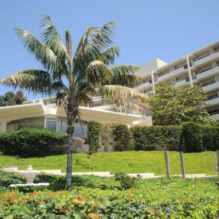 A modern hotel building surrounded by lush greenery and palm trees under a clear blue sky.