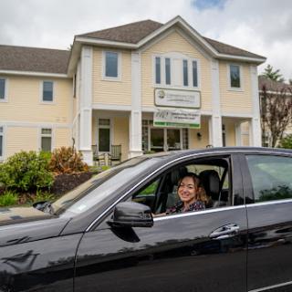 A smiling individual seated in a black car outside a charming building.