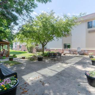 A peaceful garden patio area with benches, trees, and a building in the background.
