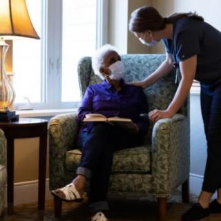A caregiver attending to an elderly person sitting in a chair, both wearing masks.